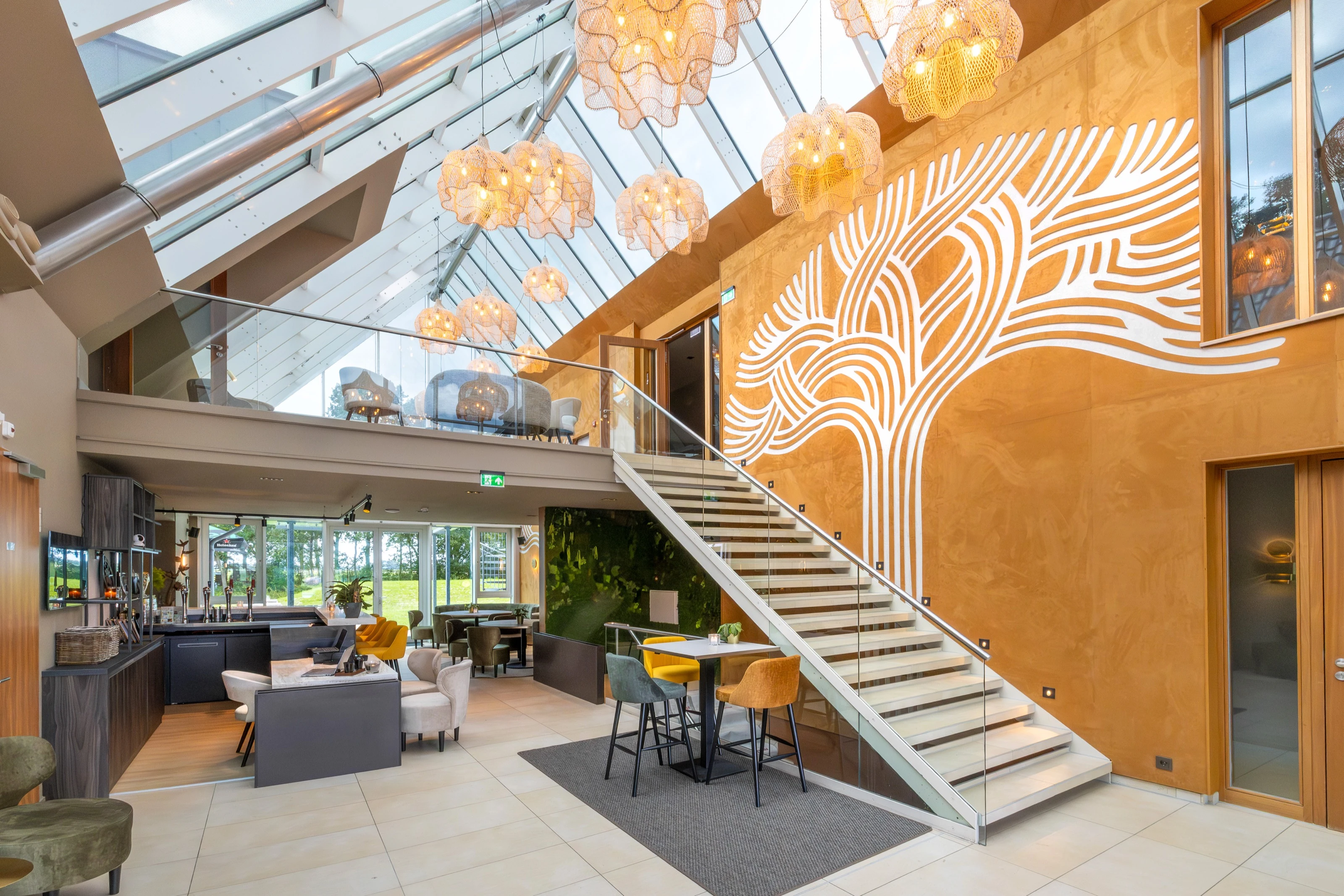 Het atrium van Cozmoss met glazen serredak, warme lampen en de boom-muurschildering boven de trap